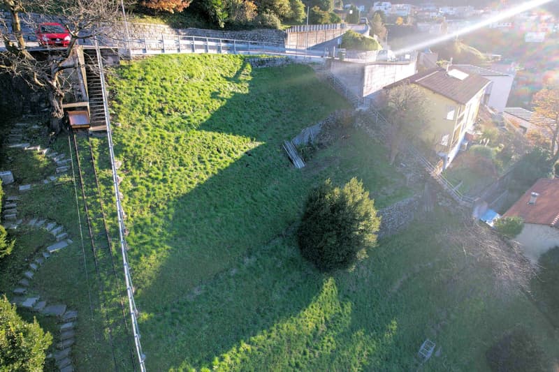 Aerial view of a grassy area with a red car parked on a bridge, a staircase leading to the bridge, and a house with a brown roof and solar panels on the side.