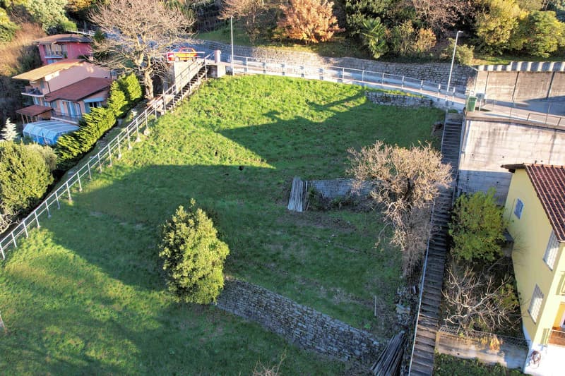 Large grassy area with a stone wall, surrounded by trees and bushes, with a house and a car in the background.