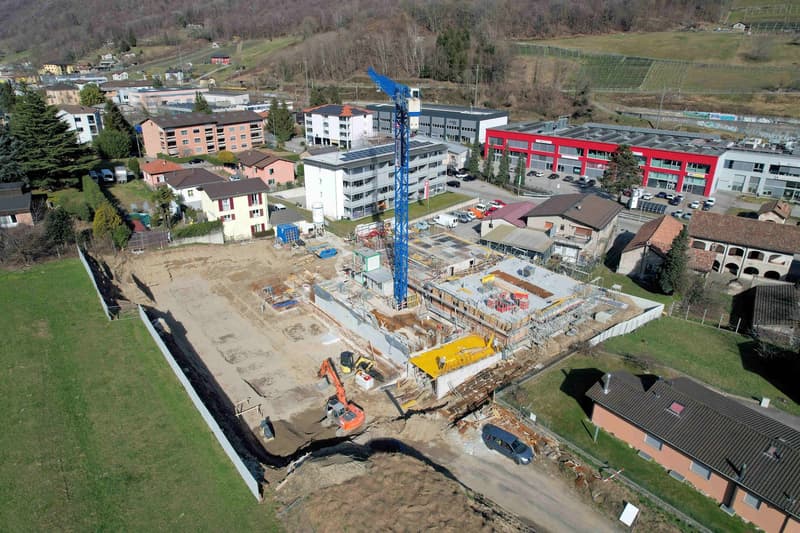 Aerial view of a construction site with a blue crane, several buildings under construction, and surrounding houses and greenery.