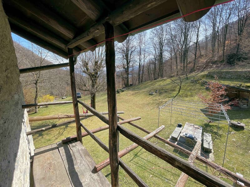 wooden balcony, wooden railings, wooden posts, grassy field, trees, car parked, wooden table, stone benches, metal structure, stone wall, mountains