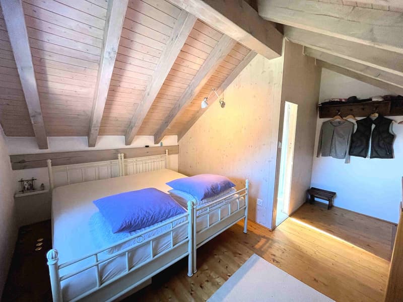 Two single beds, white bed frames, blue pillows, wooden floor, wooden ceiling, wall-mounted lamp, wooden bench, clothes hanging on the wall
