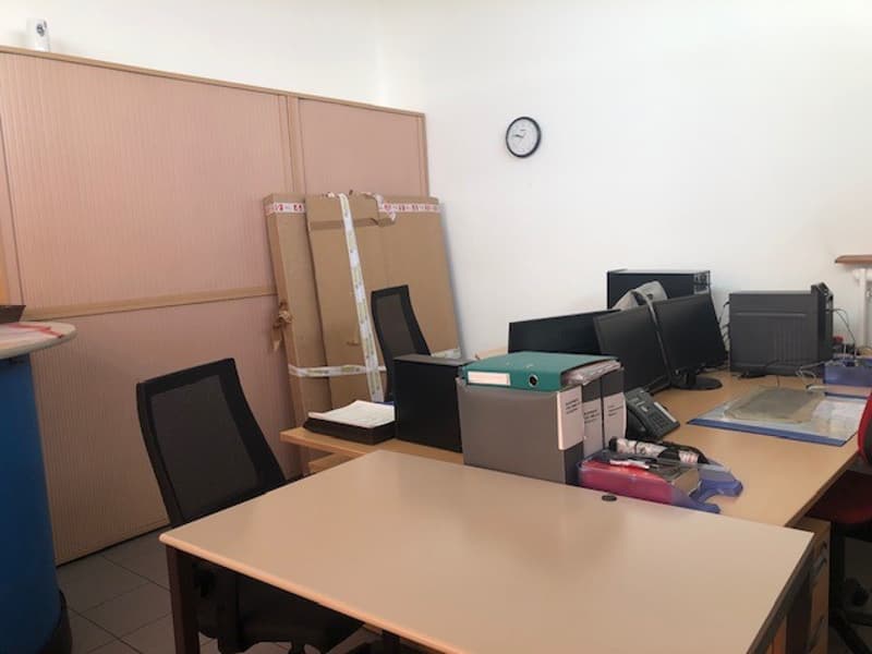 The image shows an office space with desks, computers, and office equipment. There are cardboard boxes and other items on the desks, indicating an active workspace. The room has a clock on the wall and appears to be a functional office environment.