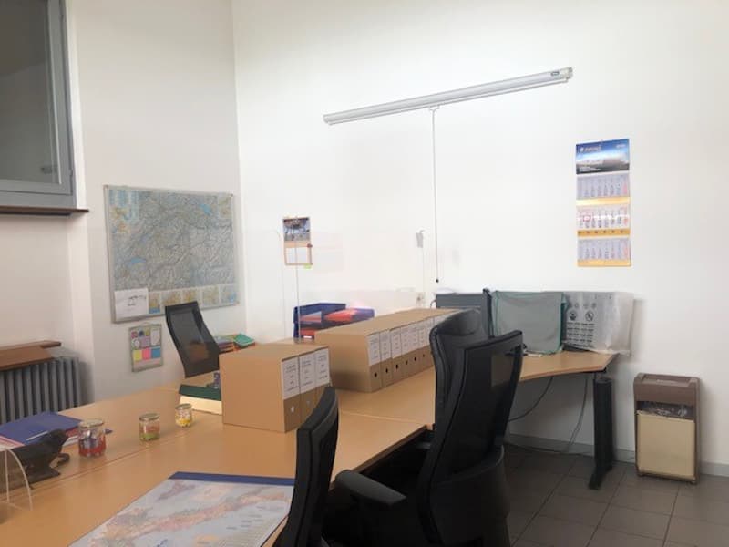 The image shows an office space with a desk, computer, and various office supplies and equipment. The room has white walls, a tiled floor, and a large window. There are also some storage boxes and a map on the wall.