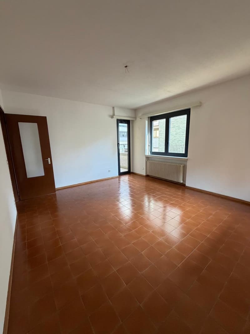 Empty room with brown tiles and white walls, door, two windows, one leading to a balcony