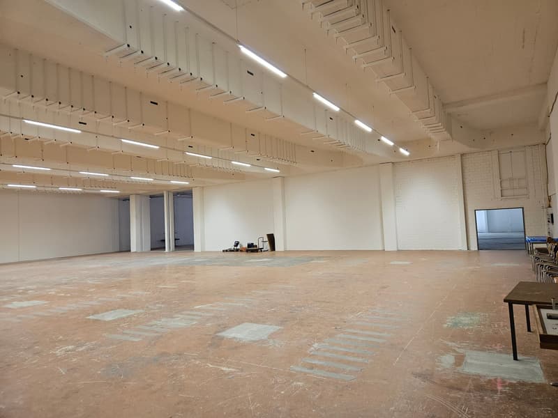 Empty room with white walls, high ceiling with many lights, open doors, concrete floor, markings on the floor.