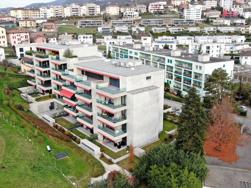 Multi-story residential complex, concrete construction, multiple balconies, landscaped gardens, several cars parked in driveway, trees in vicinity