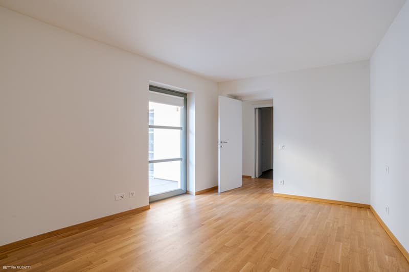 An empty room with white walls and wooden floors. The room has a door leading to another room and a glass door leading to a balcony. There is a window on the wall and power outlets.