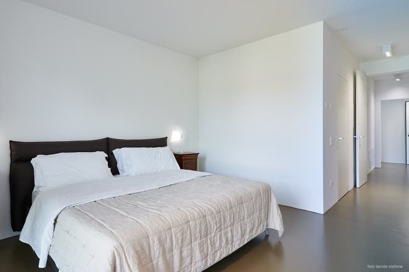 Spacious bedroom with white walls, a large bed with a gray comforter, and a wooden nightstand. The room has a minimalist and clean design.