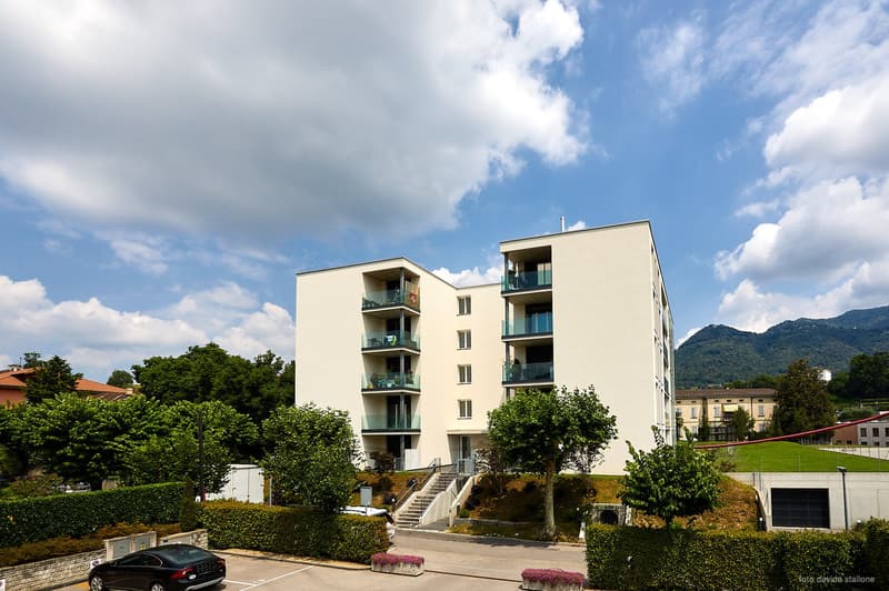 A multi-story apartment building with balconies, surrounded by trees and greenery. The building has a modern, minimalist design with clean lines and large windows. There appears to be a parking area or garage at the base of the building.