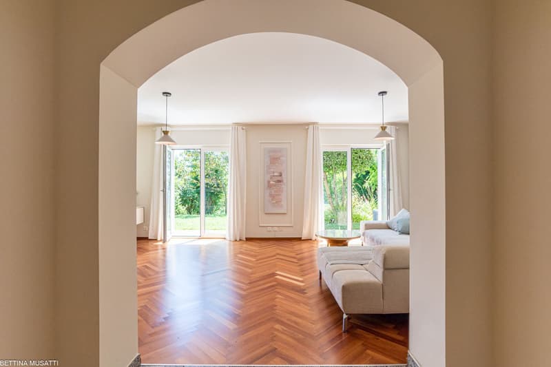 Spacious living room with large windows overlooking a garden, hardwood herringbone floor, white walls, arched entryway, pendant lighting, and a comfortable sofa