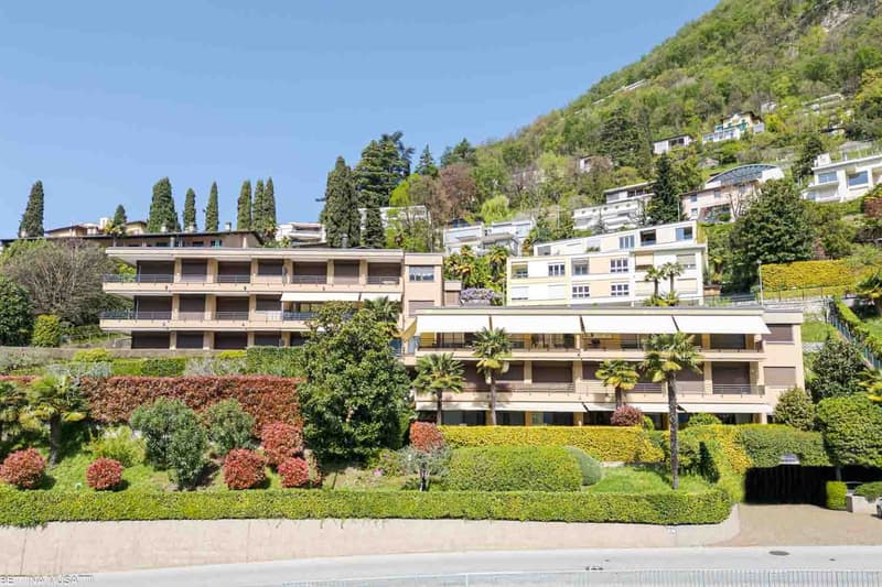 Multi-story apartment building with balconies, surrounded by lush greenery and landscaping, located on a hillside with a view of the surrounding area.