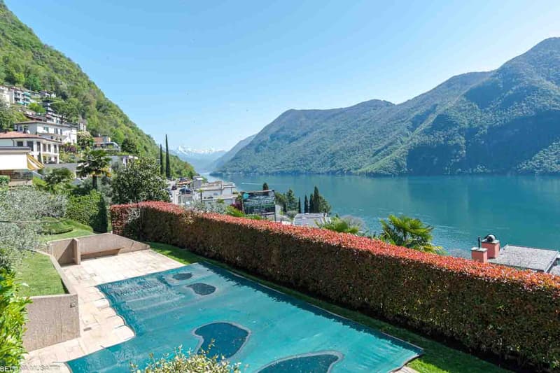 Luxury villa with a swimming pool overlooking a scenic lake surrounded by mountains. The property has a well-manicured garden with a hedge lining the edge, providing privacy. The lake and mountains in the background create a picturesque and serene setting