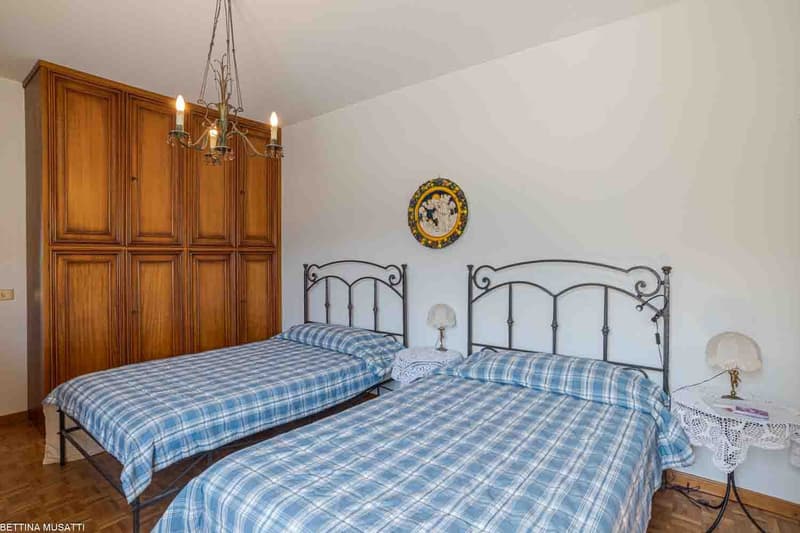 The image shows a bedroom with a wooden wardrobe, two iron-framed beds with plaid bedding, a decorative wall clock, and a table lamp. The room has a cozy and traditional feel.