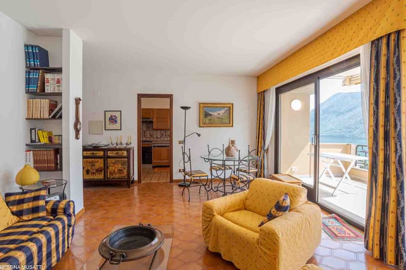Spacious living room with large windows offering a scenic view, yellow and blue color scheme, wooden furniture, bookshelves, and a balcony or terrace access