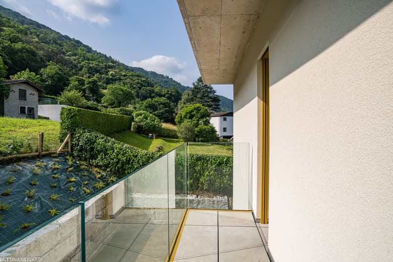 The image shows a balcony or terrace of an apartment or residential unit. The balcony has a glass railing and overlooks a lush, green landscape with mountains in the background. The building appears to be modern with a concrete ceiling and white walls. Th