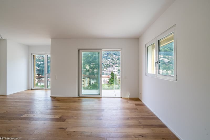 Spacious living room with large windows offering a scenic view of the surrounding landscape and buildings. The room features hardwood flooring and white walls, creating a bright and airy atmosphere.