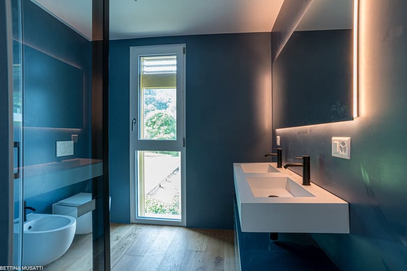 Modern bathroom with blue and dark gray walls, large window with a view, white sink vanity, and a toilet