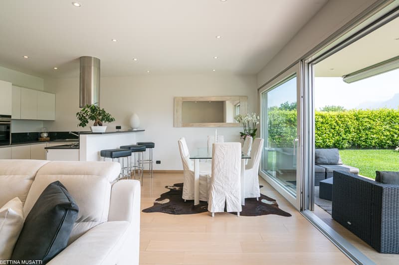 Open-concept living room and kitchen with large windows overlooking a garden, glass dining table with white chairs, beige sofa, and black rattan furniture on the patio outside
