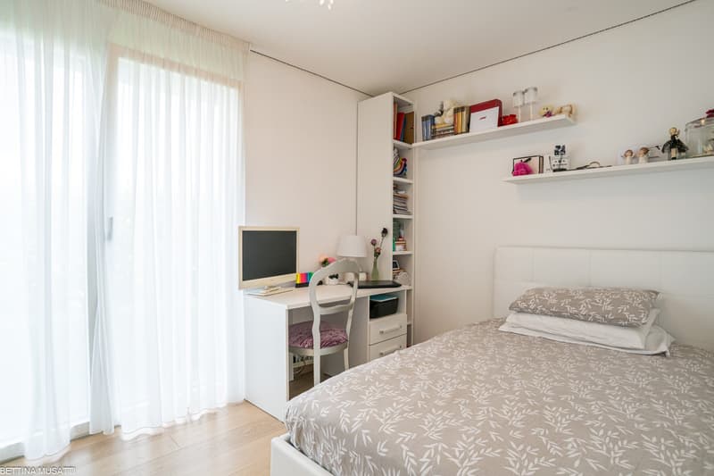 Spacious bedroom with large windows, white walls, hardwood floors, a desk with a computer, shelves with books and decor, and a patterned bedspread on the bed.
