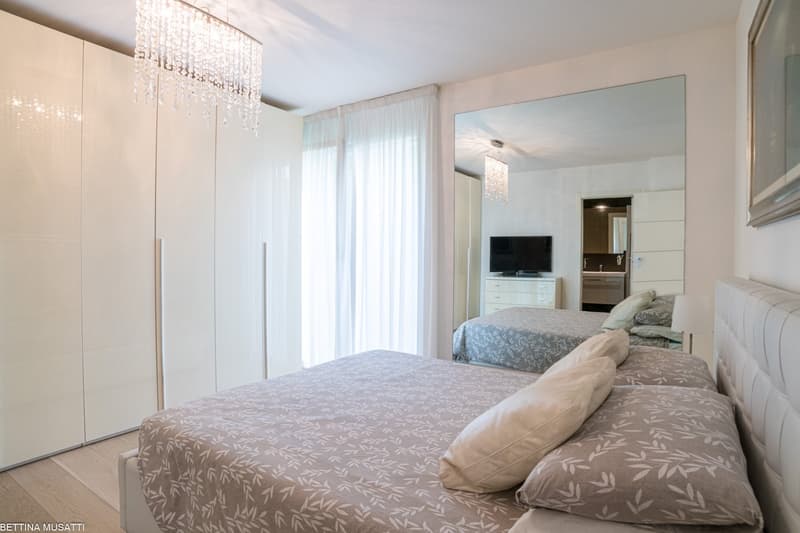 Spacious bedroom with large windows, white curtains, a crystal chandelier, a white wardrobe, a TV stand, and a king-size bed with patterned bedding.