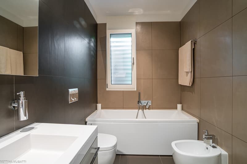 The image shows a modern and spacious bathroom with a large bathtub, a sink, and a toilet. The walls are covered with beige and dark brown tiles, creating a sleek and minimalist design. There is a window that provides natural light, and a towel rack is vi