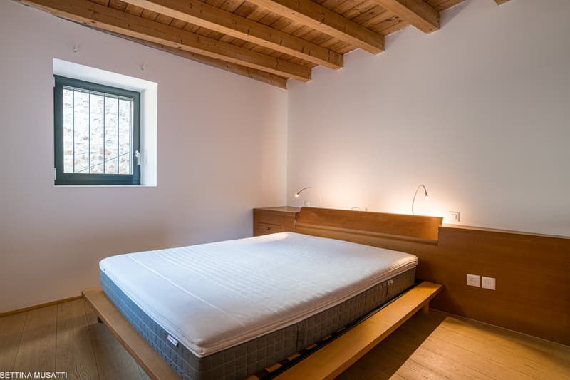 Spacious bedroom with wooden beamed ceiling, large window, wooden bed frame and nightstand, and a fireplace