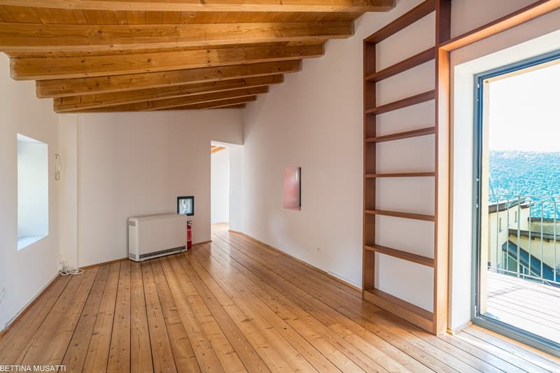 Spacious room with wooden beamed ceiling, hardwood floors, and a large window with a view. The room features built-in shelving and a small heater unit.