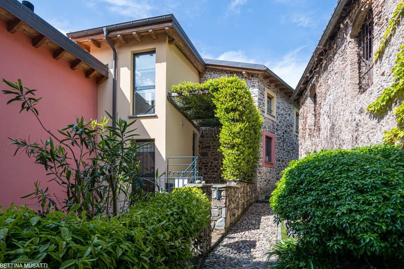Two-story house with a mix of stone and stucco exterior, large windows, balcony, and lush greenery surrounding the property. The house has a traditional architectural style with a sloped roof and wooden beams. The path leading to the house is paved with c