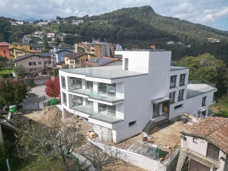 Modern white building, several floors, glass balconies, construction site in front, surrounded by houses and a mountain