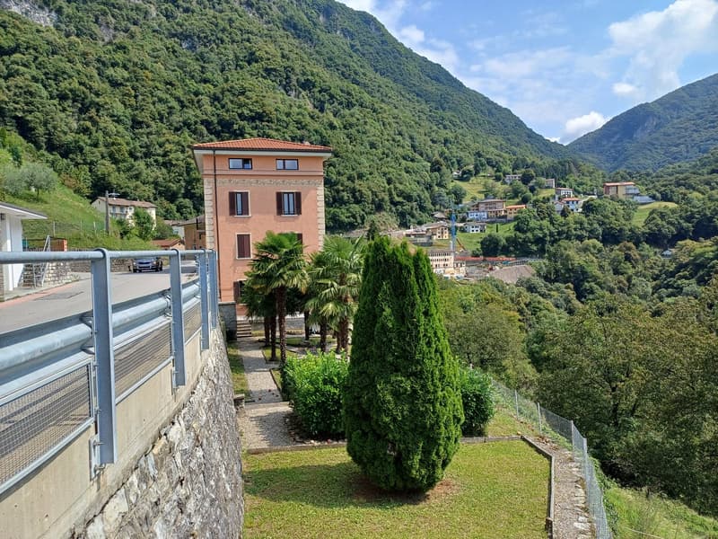 A two-story brick building, set in a hillside community with steep terrain, featuring several windows and a tiled roof. It is surrounded by lush greenery, including multiple trees and bushes, and is positioned against a mountainous backdrop. A car is park