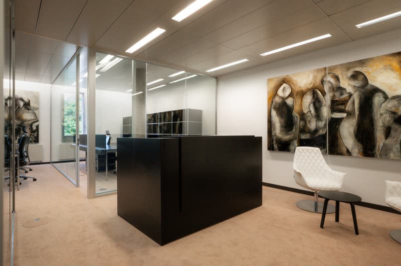 Contemporary office space with large glass walls, minimalist furniture, black central desk, modern paintings on the wall.