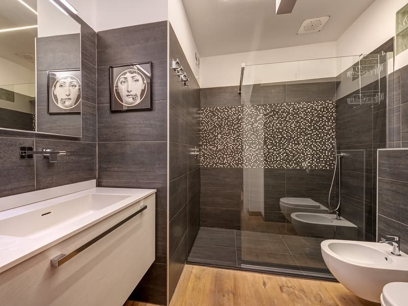 Modern bathroom with gray tiled walls, wooden floor, large mirror, white sink, large glass shower enclosure, white toilet, shower rack