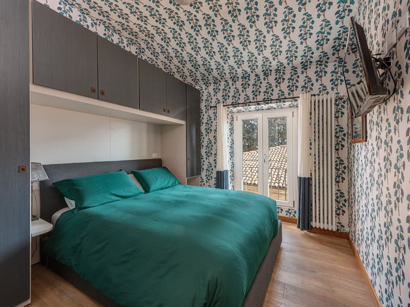 wooden floor, built-in cabinets, bed with green sheets, nightstand with lamp, TV mounted on the wall, balcony view