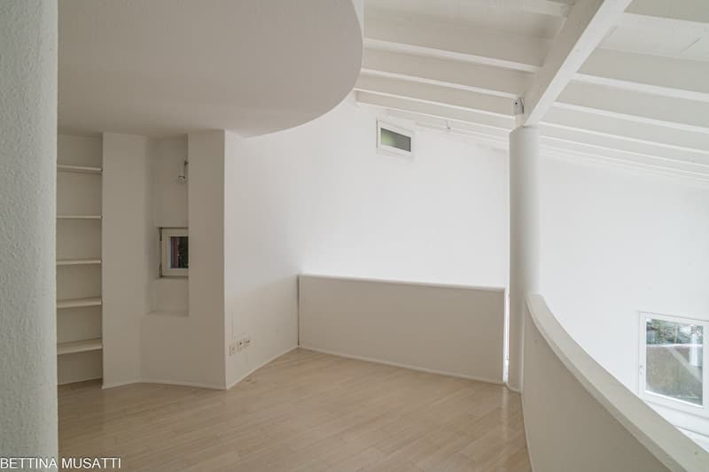 Empty room with white walls, wooden floor, built-in shelves, round arched section, square window.
