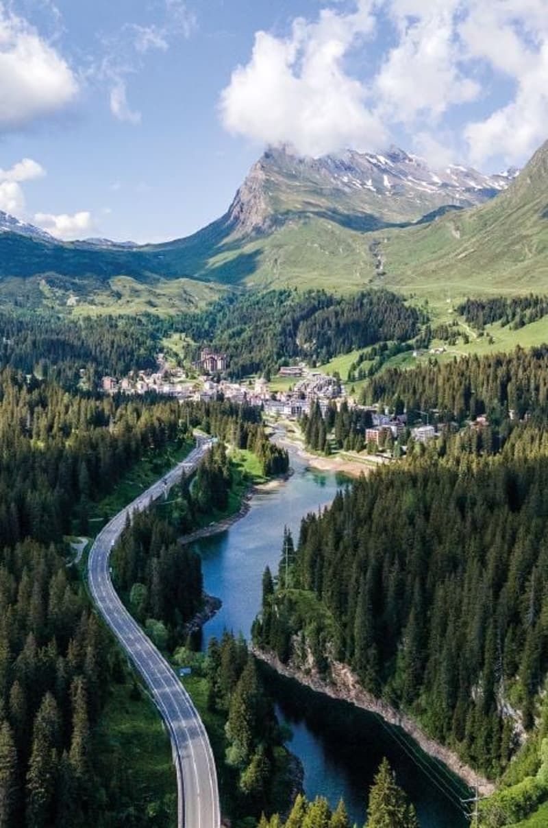 Aerial view of a highway running alongside a river, surrounded by mountains, forests, and a town