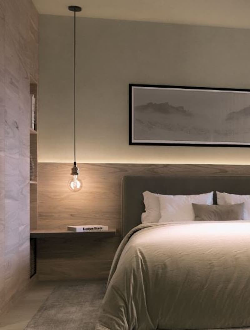 A bedroom with a large bed, wooden wall, ceiling pendant light, a framed photo on the wall, a wooden shelf with a book, and a rug.
