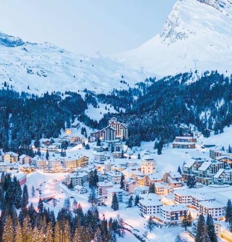 Snow-covered mountainside town, several buildings with mountain view, pine trees, ski slope