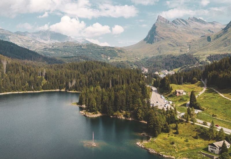 hilly landscape with large lake, mountains, forest, a single house, a small village, a road with cars, a red car on side