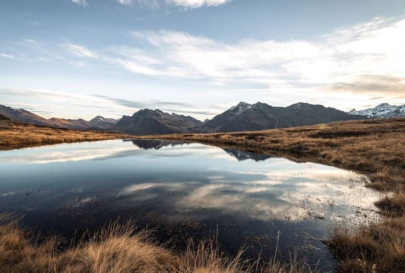 a pond with grass on both sides, mountains in the background, clouds in the sky