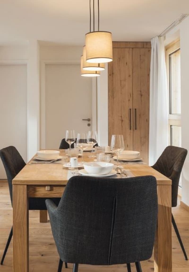 Wooden dining table, black chairs, wooden cabinet, chandelier, wine glasses, plates, bowls, white walls, windows