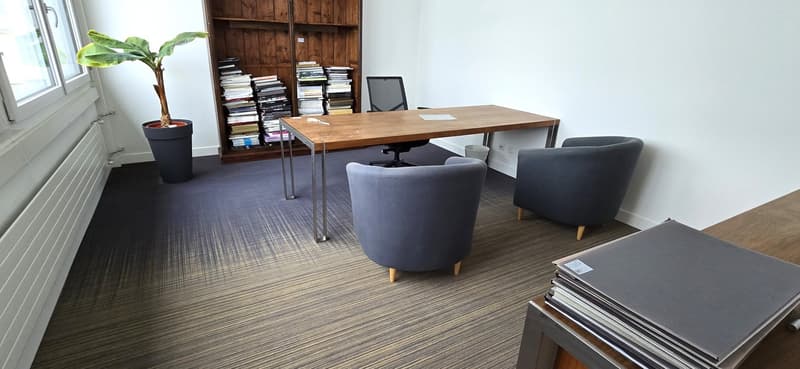 A modern office with a wooden desk, two black chairs, a bookshelf full of books, a potted plant, a radiator and gray office chair.