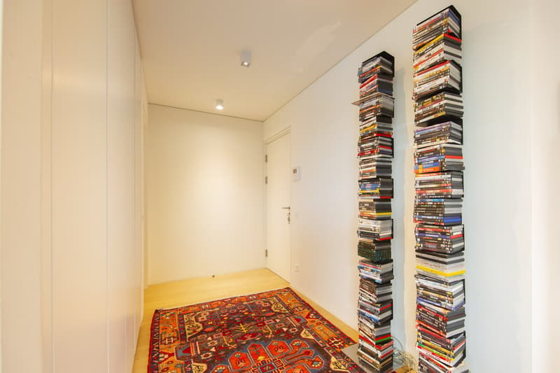 Empty hallway with white walls, wooden floor, two stacks of DVDs, a rug, and a door.