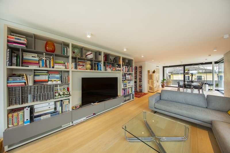 Living room with a large bookshelf, a flat screen TV, a gray couch, a glass table, and a glass door leading to a balcony.