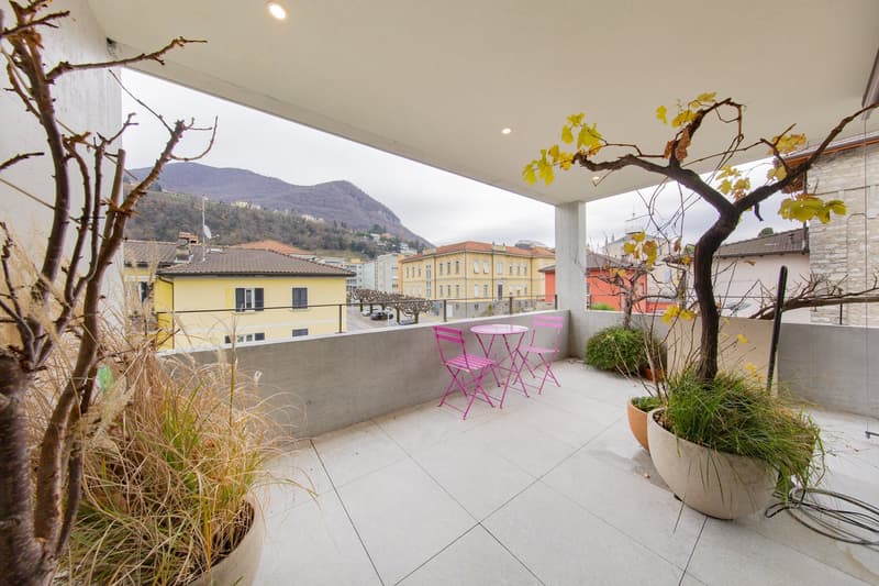 Balcony with tiled floor, metal railing, pink chairs, small table, potted plants, distant view of buildings and mountains