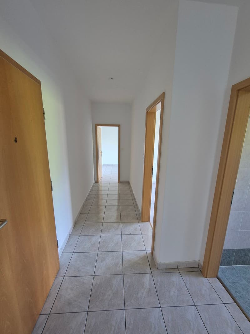 The image shows a long, tiled hallway with white walls and wooden doors leading to other rooms. The hallway has a clean, minimalist appearance with a simple and functional design.