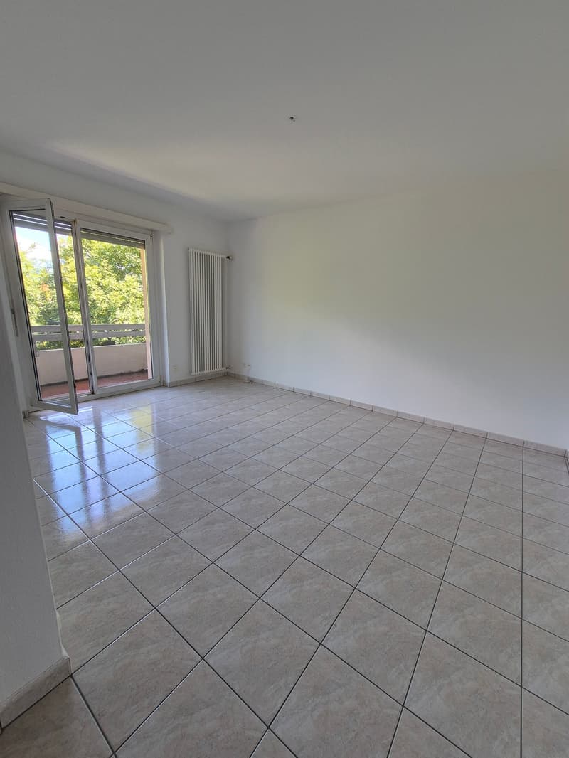 The image shows a spacious room with white walls and a tiled floor. The room has a large sliding glass door that leads to a balcony or terrace, providing natural light and a view of the outdoors. The room appears to be empty, with no furniture or decor vi