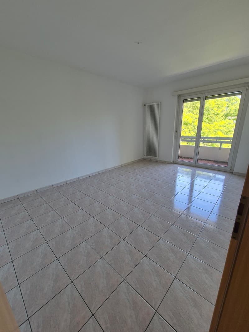 Spacious room with large windows, tiled flooring, and a balcony or terrace overlooking a lush, green outdoor area.