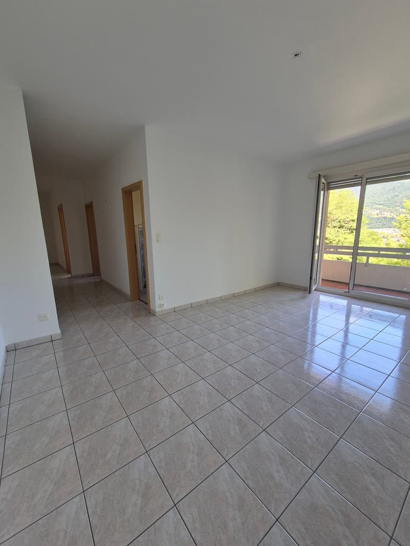 The image shows a spacious room with a large window overlooking a scenic outdoor view. The room has a tiled floor in a light gray color, and the walls are painted in a neutral tone. There are two wooden doors visible, likely leading to other rooms or area