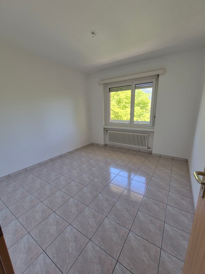 Empty room with white walls, large window overlooking greenery, and gray tile flooring.