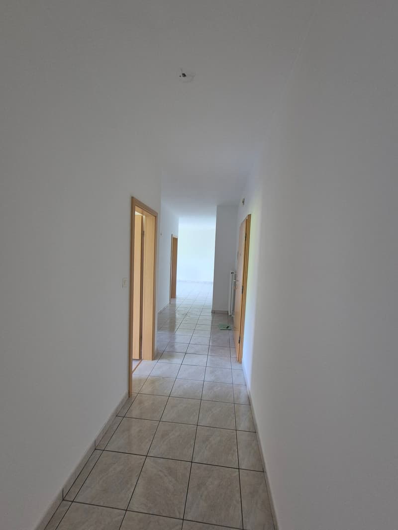 The image shows a long, narrow hallway with white walls and a tiled floor. There are two wooden doors visible, one on the left and one on the right, leading to other rooms. The hallway appears to be well-lit and has a clean, minimalist design.